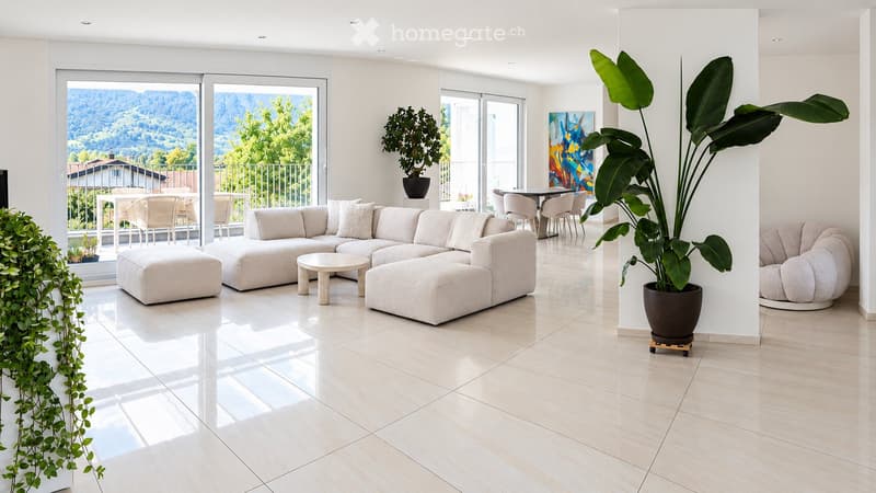 Modern living room with large windows offering a scenic view, featuring a light-colored sectional sofa, coffee table, ottoman, dining area with chairs and table, and potted plants.