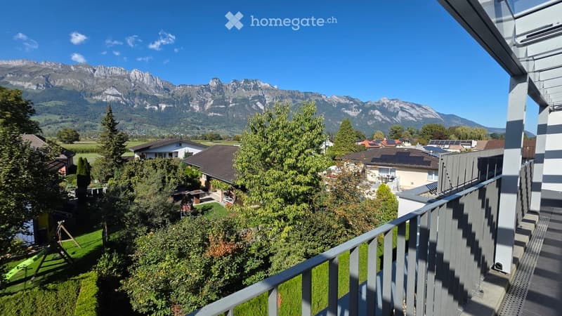 Balcony overlooking a scenic valley with mountains in the background, surrounded by trees and houses.
