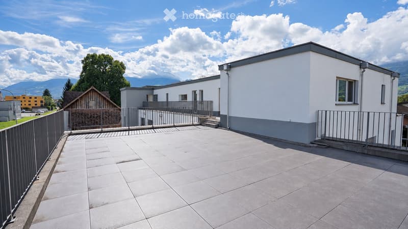 A spacious, tiled balcony or terrace area with a metal railing, steps leading up to a building entrance, and a view of surrounding greenery and mountains.