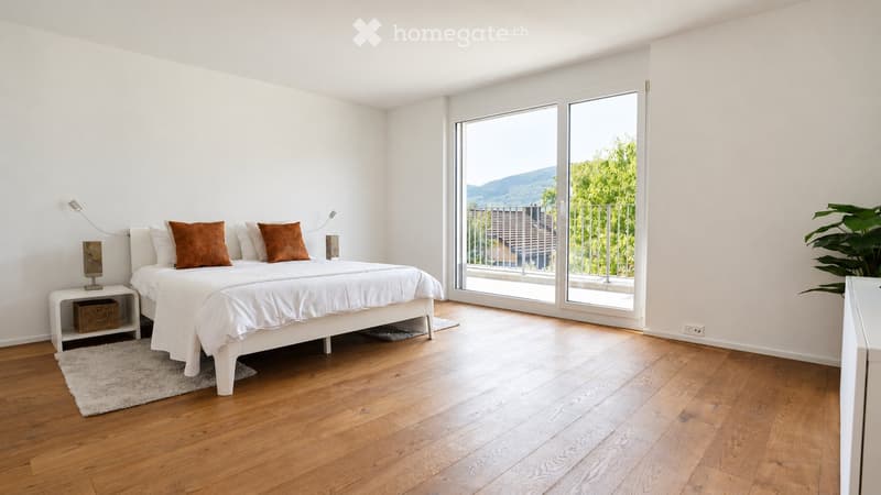 Bright bedroom with wooden flooring, large window offering a scenic view, a white bed with brown pillows, a nightstand with a lamp, and a small plant near the window.