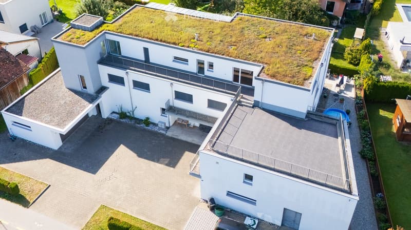 Aerial view of a modern, white house with a green roof, multiple balconies, and a swimming pool in the backyard.