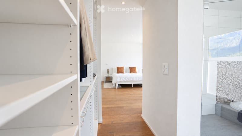 Modern bedroom with a white bed, nightstand, and two brown pillows. The room features wooden flooring, a white wardrobe, and an adjacent bathroom with a bathtub and a large window offering a scenic view.