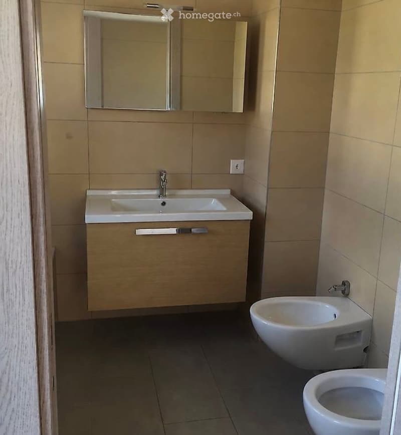 Modern bathroom with beige tiles, a white sink with a mirror above, and a toilet and bidet below.