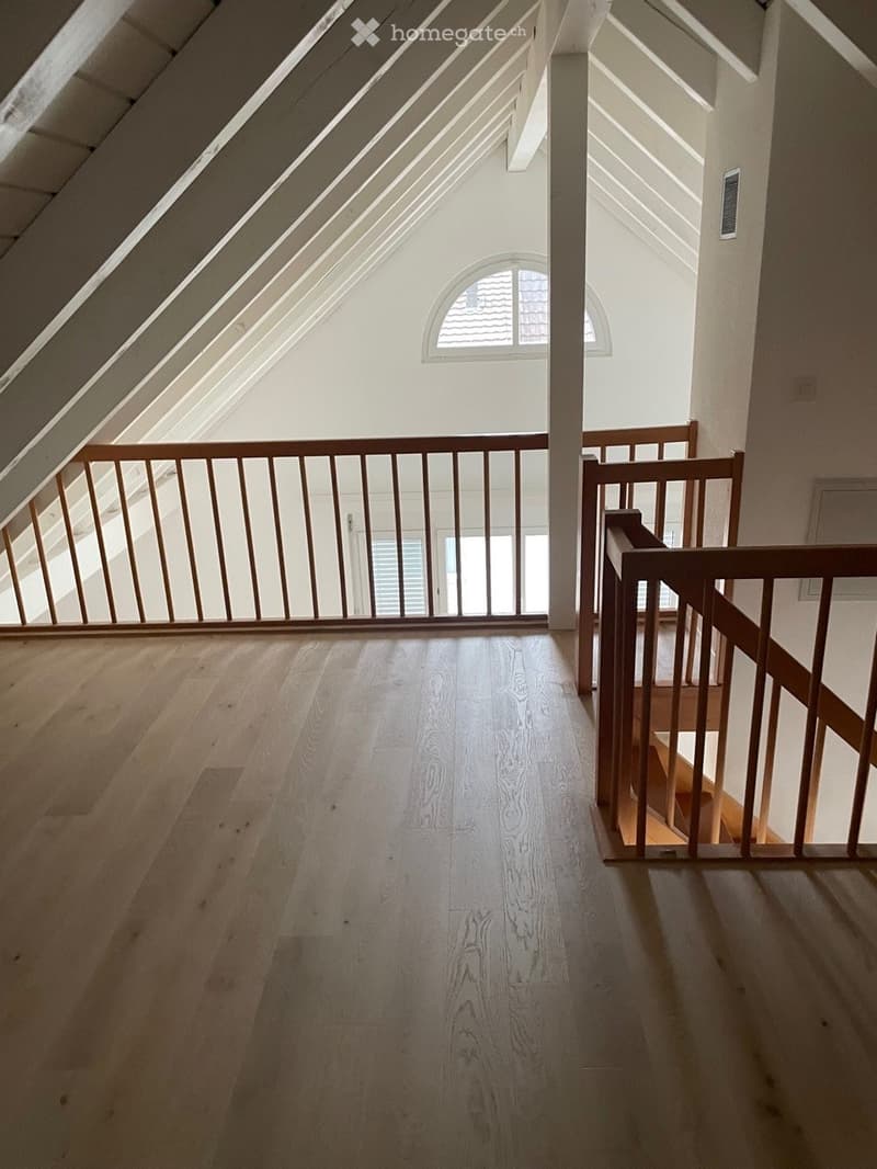 Empty attic space with wooden flooring, exposed wooden beams, and a wooden railing leading to a staircase. Natural light streams in from a skylight and a window.
