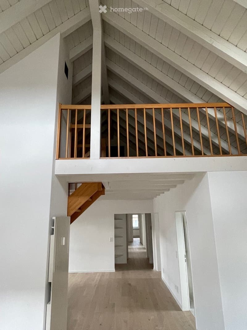 Bright, open hallway with white walls, wooden staircase leading to an upper level, and visible attic space with exposed wooden beams and slanted ceilings.