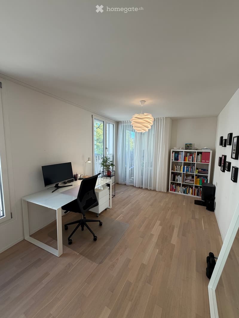 Modern, well-lit room with wooden flooring, a white desk, ergonomic chair, large screen, bookshelf, and a hanging lamp.