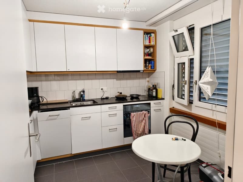 A compact kitchen with white cabinets, a black countertop, and a small dining area featuring a round table and chair. The kitchen includes a sink, oven, coffee maker, and various small appliances and utensils.