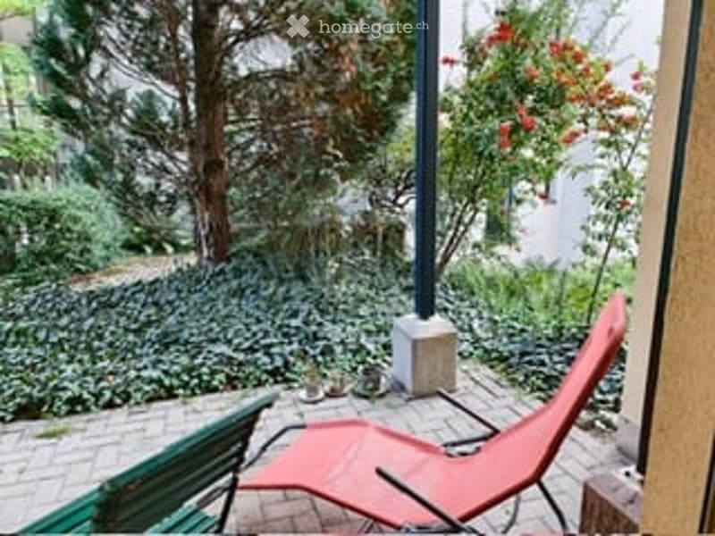 Balcony with a red lounge chair and a green bench, overlooking a garden with trees and shrubs.