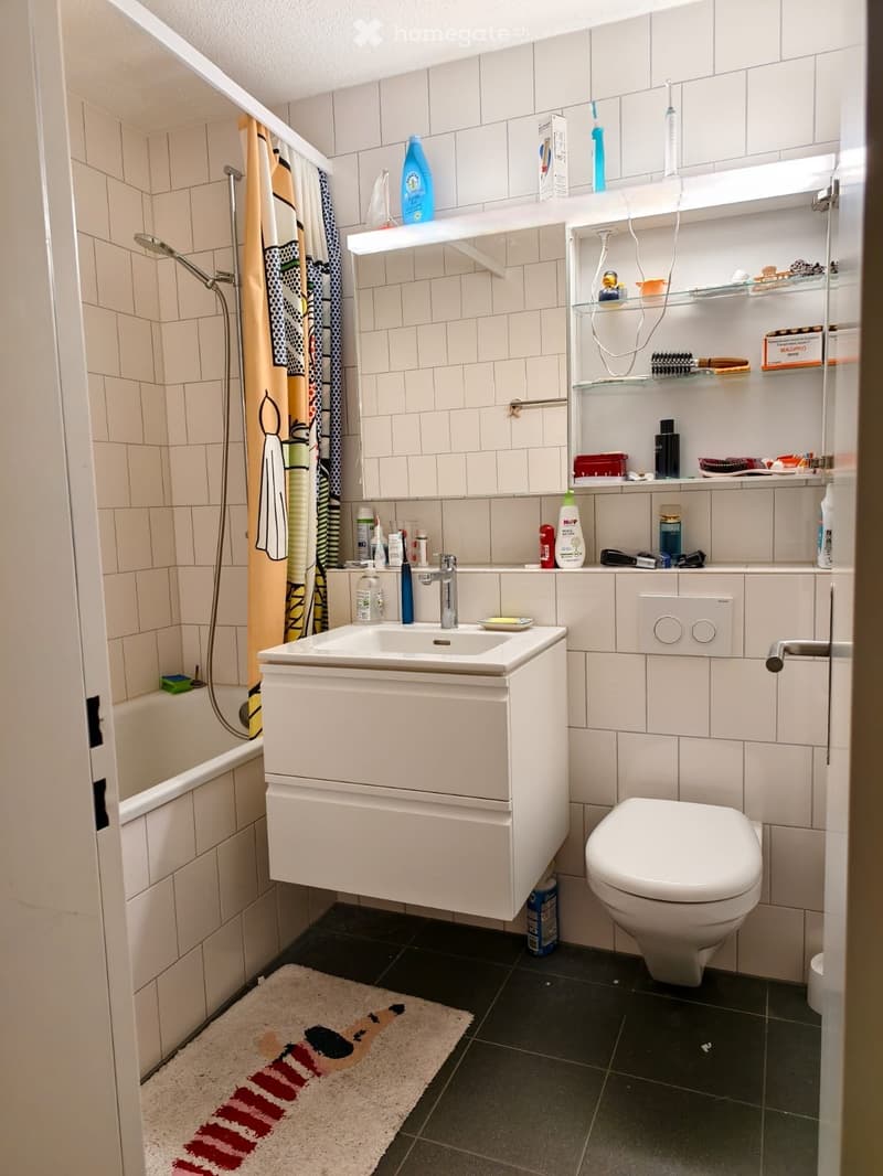 Modern bathroom with white tiles, a sink with a mirror, a toilet, a bathtub with a shower, shelves with toiletries, and a colorful rug on the floor.