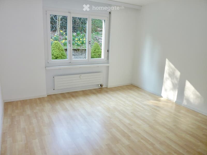 Empty room with light wooden flooring, white walls, and large windows showing a garden view. No furniture or equipment present.