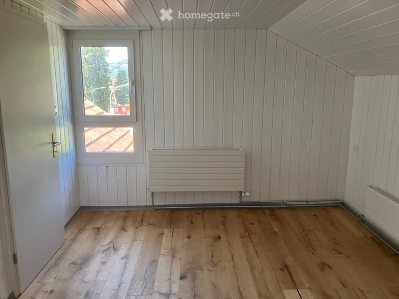 Empty room with wooden flooring, white paneled walls, and radiators. Features a window with a view of the outside and a door leading to another space.
