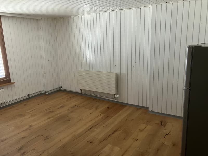 Empty room with wooden flooring, white paneled walls, radiator, and a window with wooden frame.