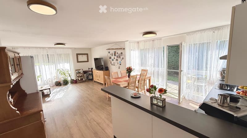 Open-plan living and dining area with wooden flooring, a kitchen counter, a dining table with chairs, a television, and a refrigerator. The space is well-lit with large windows and sheer curtains, and includes decorative elements like flower vases and a c