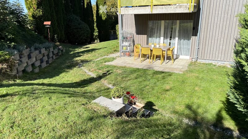 Well-maintained garden with a raised patio area featuring a dining set, surrounded by lush greenery and a modern house with a balcony.