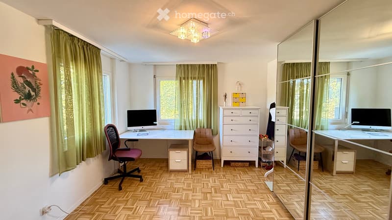A well-lit room with wooden flooring, a large mirror, and a desk setup with a computer and chair. The room features a drawer unit, a painting on the wall, and green curtains on the windows.