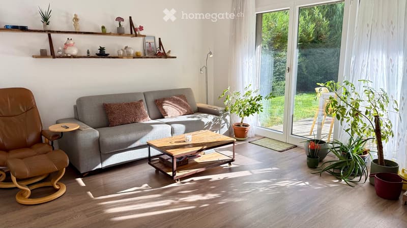 Modern living room with a grey sofa, wooden coffee table, leather armchair, and potted plants near large windows.