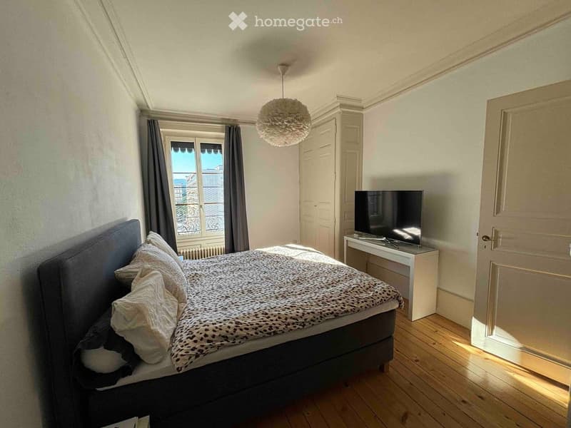 A well-lit bedroom with wooden flooring, a large window with curtains, a bed with pillows and a patterned blanket, a television on a stand, and a hanging light fixture.