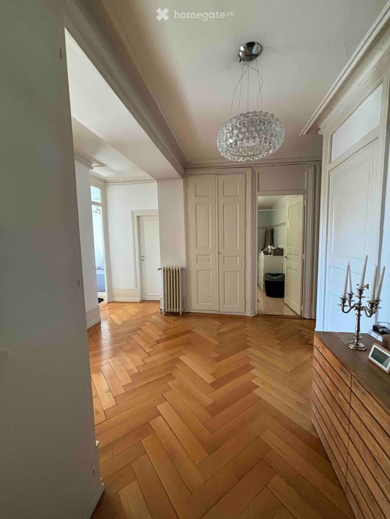 A spacious hallway with herringbone wooden flooring, white walls, and a modern chandelier. Features include a radiator, a cabinet, a candelabra, and a mirror reflecting part of the adjacent room.
