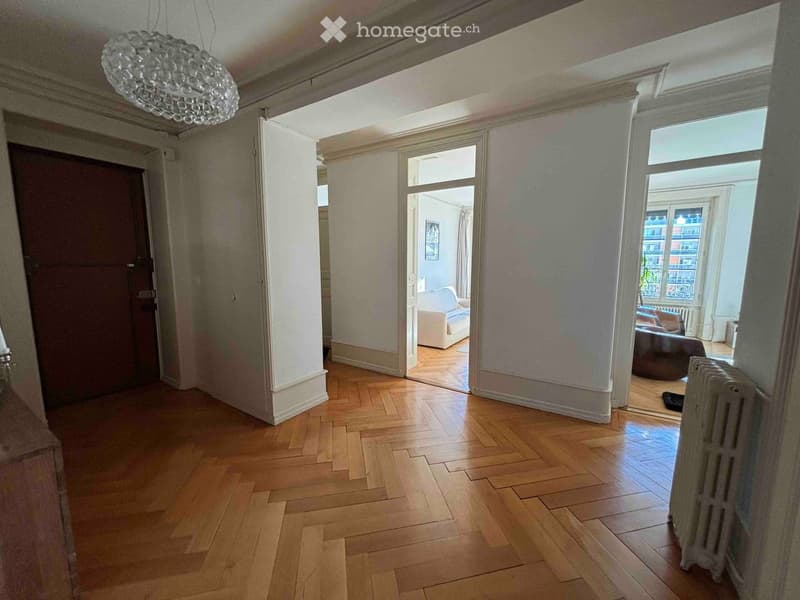 Spacious hallway with wooden flooring, large central column, and open doorways leading to adjacent rooms. Visible features include a chandelier and radiator.