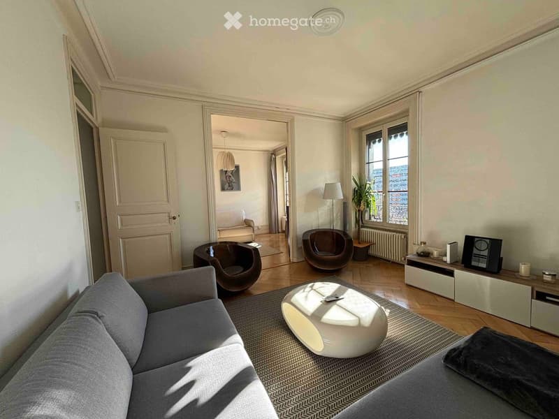 Modern living room with grey sofa, white coffee table, two brown chairs, a small table, a lamp, a TV screen, and a plant near the window.