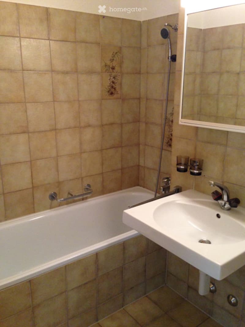 Bathroom with beige tiled walls and floor, a white bathtub, a white sink with a chrome faucet, a mirror above the sink, and a showerhead with a handheld shower attachment.