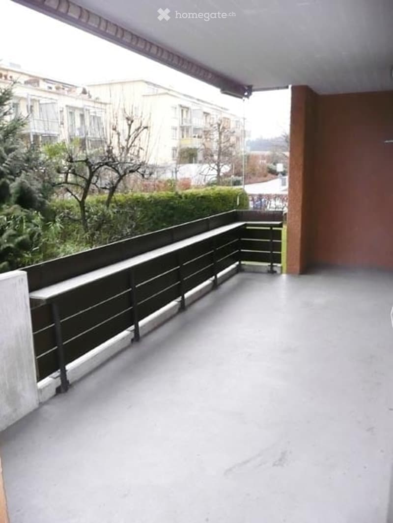 Balcony with metal railing, concrete floor, and outdoor seating area overlooking a landscaped garden and neighboring buildings.