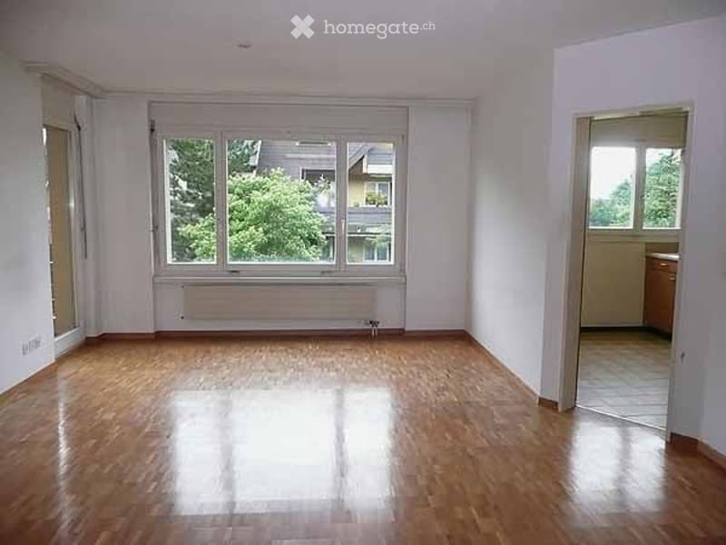 Empty room with wooden flooring, large window with white frame, radiator below window, and an open doorway leading to another room.
