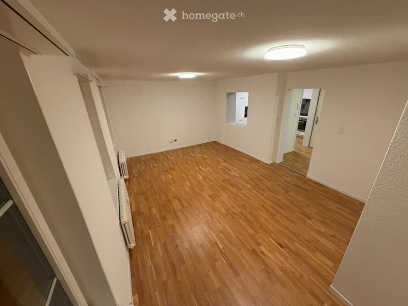 Empty hallway with wooden flooring, white walls, and recessed lighting. A small window and a radiator are visible on one wall, with an open doorway leading to another room.