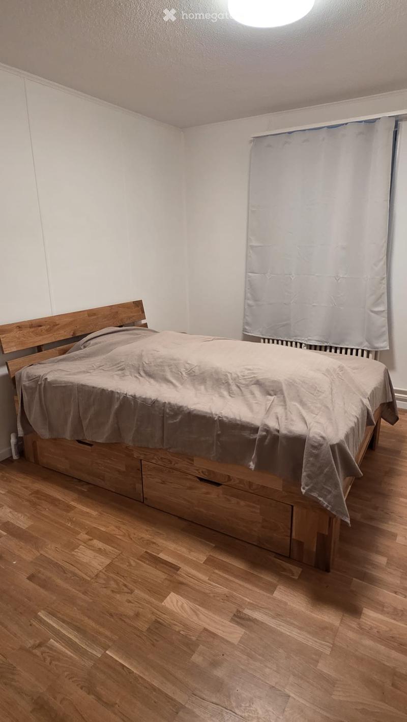 A small bedroom with a wooden bed, a grey duvet, and a curtain on the window. The floor is made of light wooden planks.