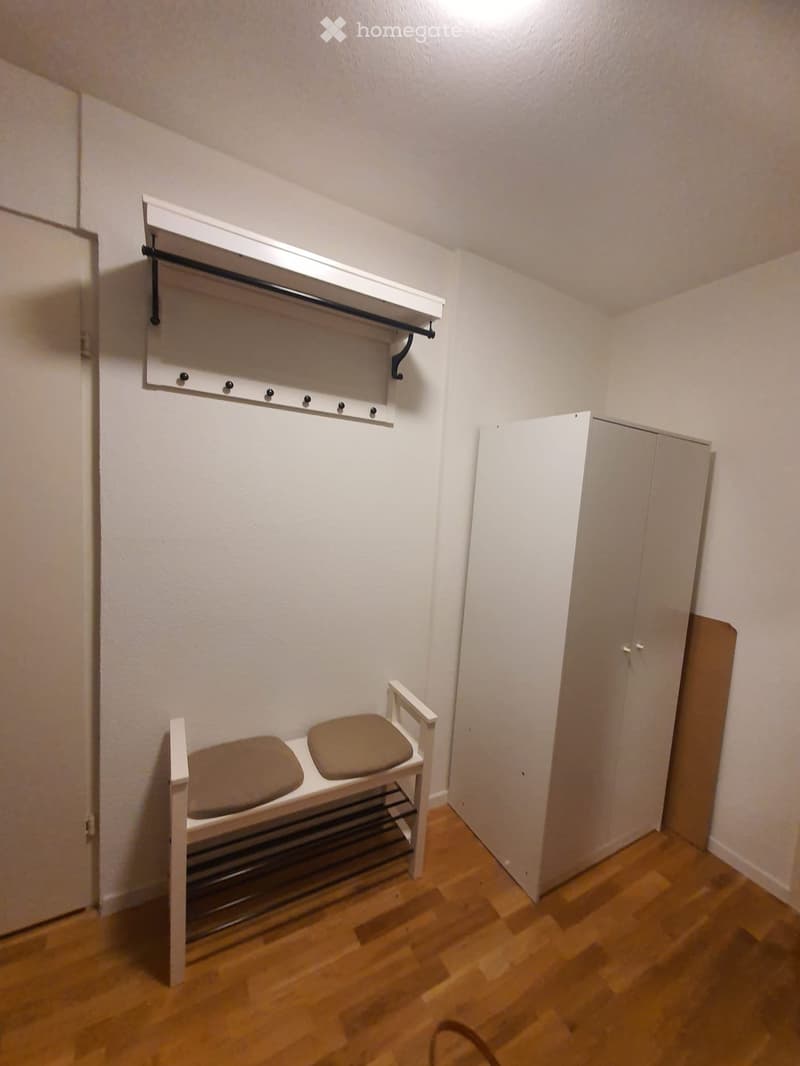 A small, well-lit hallway or entryway with a wooden floor, a white bench with two seats, a coat rack, and a white wardrobe.