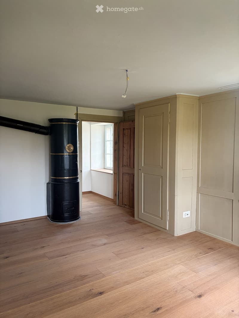 Empty room with wooden flooring, light-colored walls, and a black wood-burning stove. There is a doorway leading to another room with a window and a small bench.