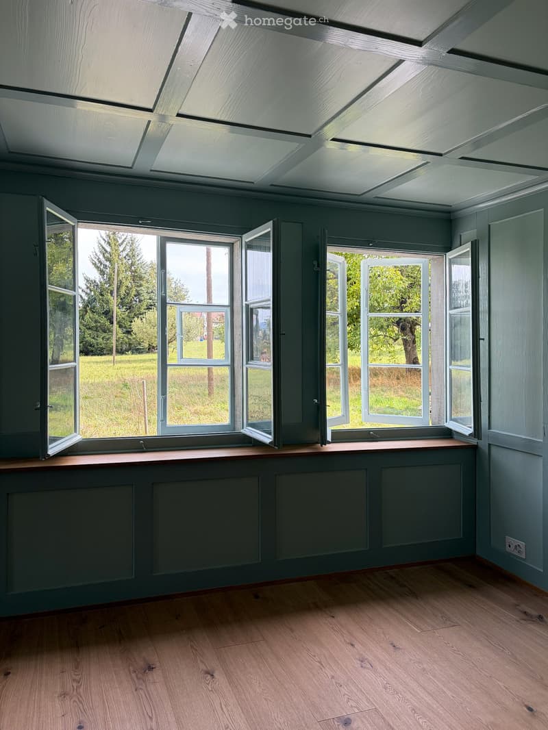 Empty room with wooden flooring, grey walls, large windows with open shutters, and a skylight ceiling.