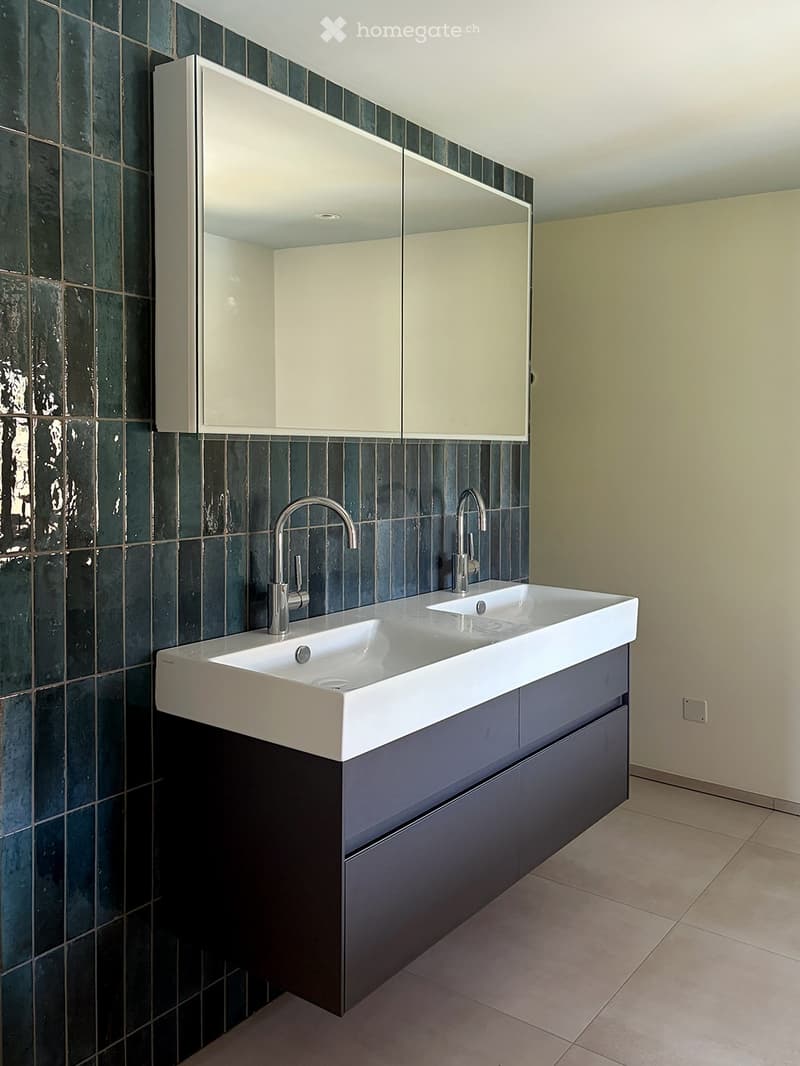 Modern bathroom with a double sink, sleek faucets, a large mirrored cabinet, and dark green tiled walls.