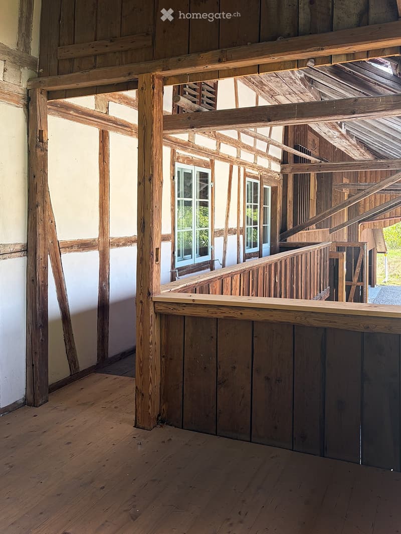 Empty wooden room with exposed beams, wooden floor, and a staircase leading to an upper level. The walls are partially finished with wooden paneling and plaster.