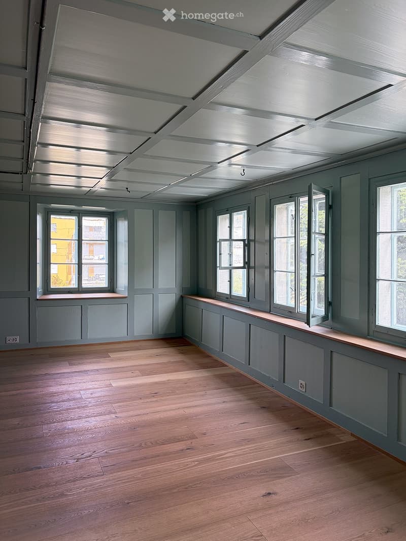 Empty room with wooden flooring, grey walls, and multiple windows with open shutters. The ceiling features exposed beams and skylights.