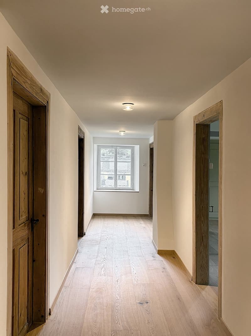 Empty hallway with light wooden flooring, white walls, and recessed lighting. Features a window at the far end and two wooden doors on either side.