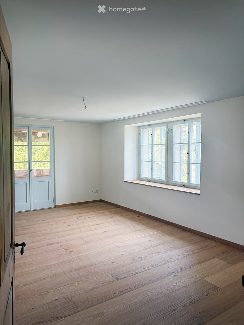 Empty room with light grey walls, wooden flooring, and large windows with white frames. A door with glass panels is visible on the left side.