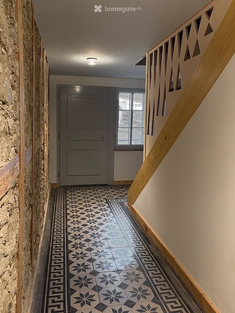Hallway with patterned tile floor, wooden staircase railing, and a closed door at the end.