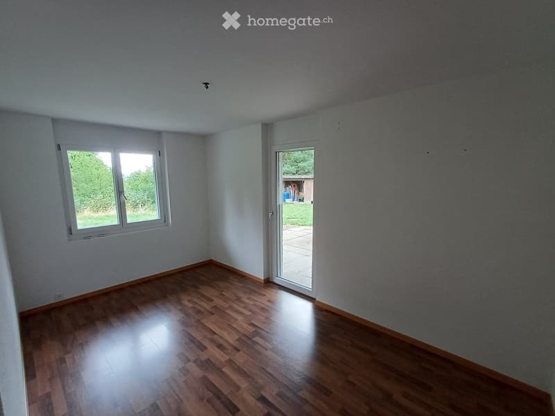 Empty room with wooden flooring, white walls, and a large window with a view of greenery. A sliding glass door leads to an outdoor patio.