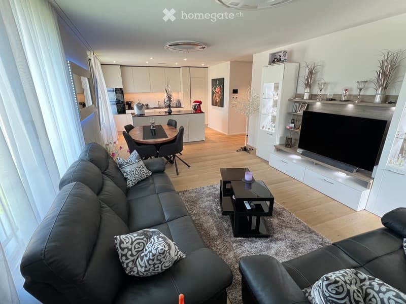 Modern living room with a black leather sofa, coffee table, TV, dining area with a round table and chairs, and a kitchen area with white cabinets and a kitchen island.