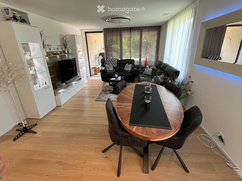 Modern living and dining area with hardwood flooring, large windows, a black leather sofa, a round wooden dining table with four chairs, a TV on a white cabinet, and decorative vases.