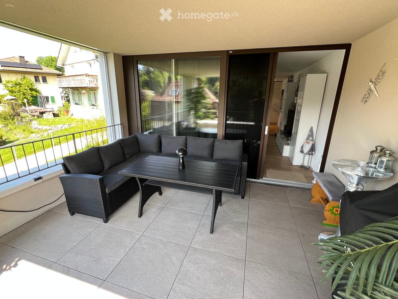 Balcony with a grey sectional sofa, black coffee table, wicker chair, small lamp, and decorative items including a small stool and a glass table with lanterns.