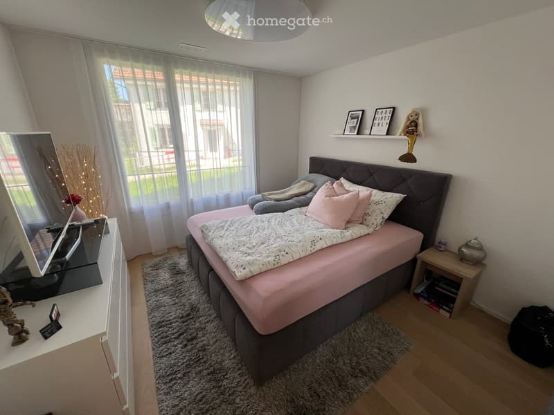 Cozy bedroom with a double bed, nightstand, mirror, and decorative shelf. The room features a large window with sheer curtains and a view of the outside.