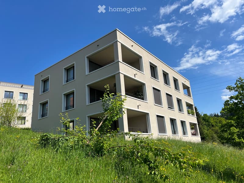 Modern multi-story building with large windows and balconies, surrounded by greenery and grass.