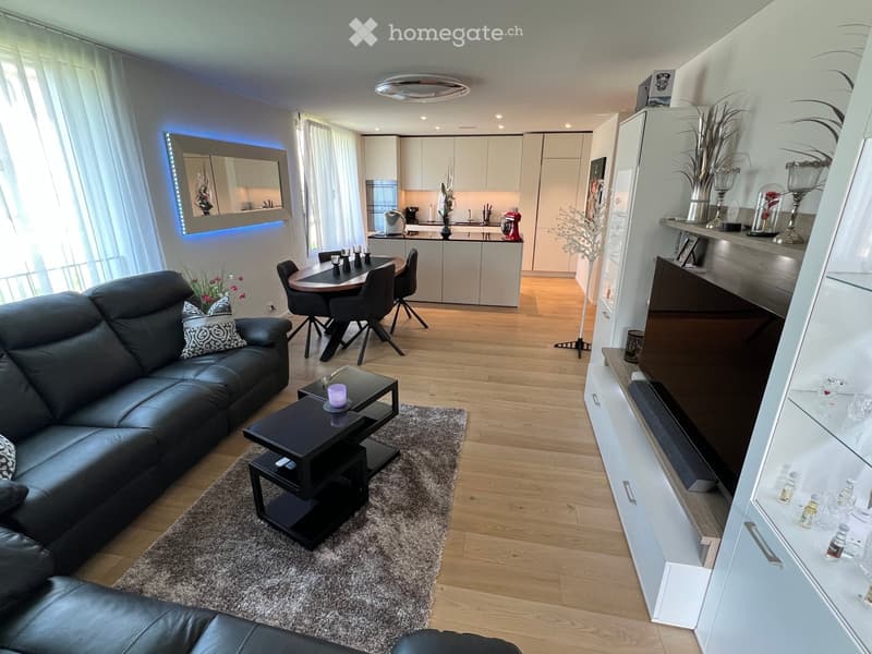 Modern living and dining area with a black leather sofa, coffee table, side table, dining table with chairs, and a kitchen area with white cabinets and appliances.