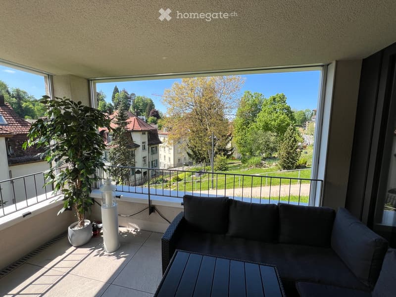 Balcony with a view of a green area, featuring a grey sofa, a small table, and a potted plant.
