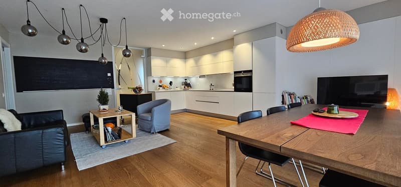 Open-plan living and dining area with modern white kitchen cabinets, a large wooden dining table, a sofa, chairs, pendant lights, and a screen.