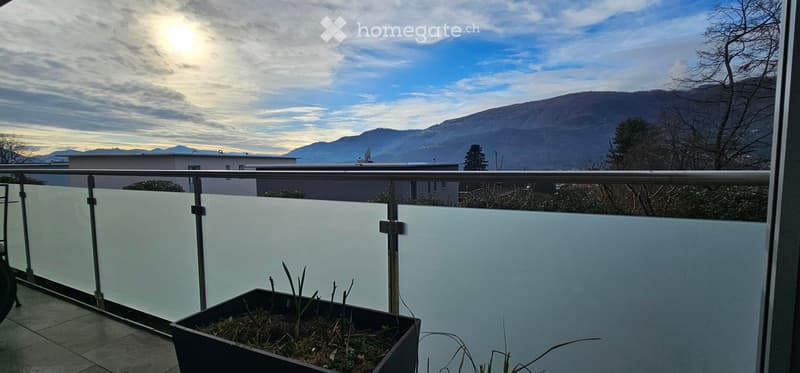 Balcony with glass railing, tiled floor, and a small planter with plants, overlooking a scenic mountain view.