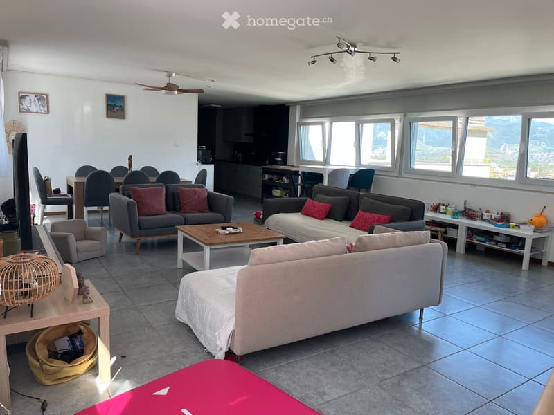 Open-plan living and dining area with large windows offering a scenic view, featuring a grey sectional sofa, wooden coffee table, dining table with chairs, and a television.