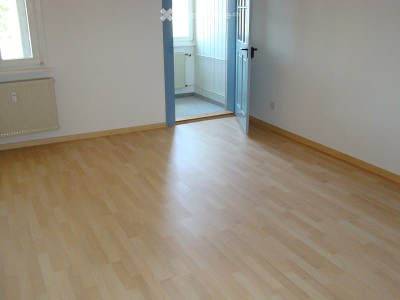 Empty room with light wooden flooring, white walls, and a radiator on the left side. A doorway leads to another room with similar flooring.